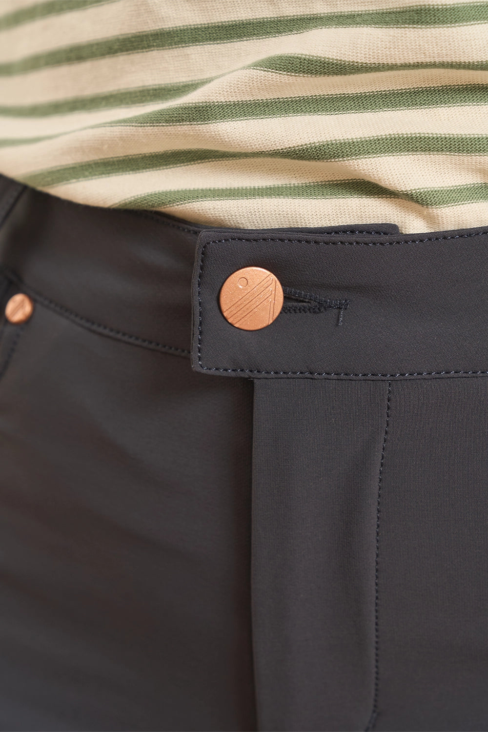 Close-up of dark charcoal gray trousers featuring a distinctive buttoned waistband and a striped knit top in beige and green visible at the top.
