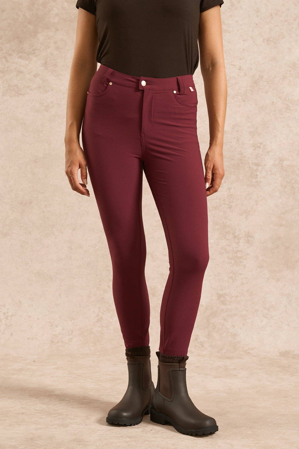 A model wearing high-waisted, slim-fit burgundy leggings paired with a black top and black rubber boots, standing against a textured beige studio backdrop.