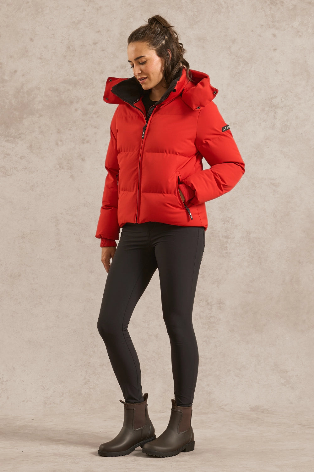 A woman modeling a bright red hooded puffer jacket, paired with black leggings and dark brown ankle boots, standing against a textured beige studio backdrop.