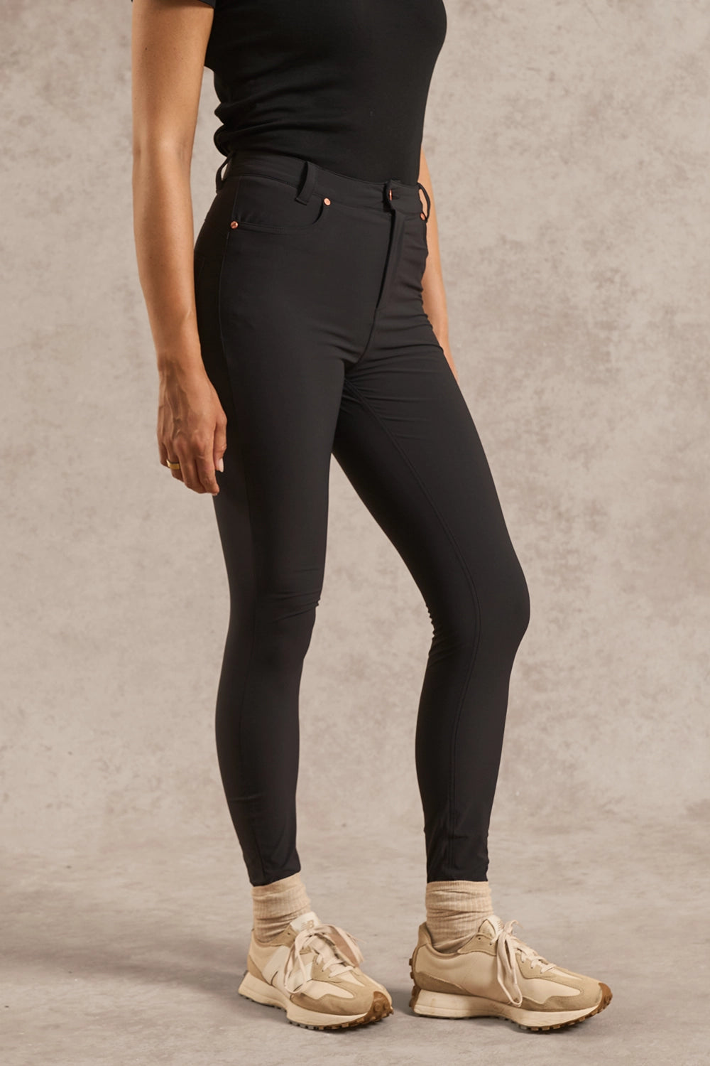 A model wearing black high-waisted skinny jeans, a black top, beige socks, and beige sneakers, standing against a textured beige studio backdrop.