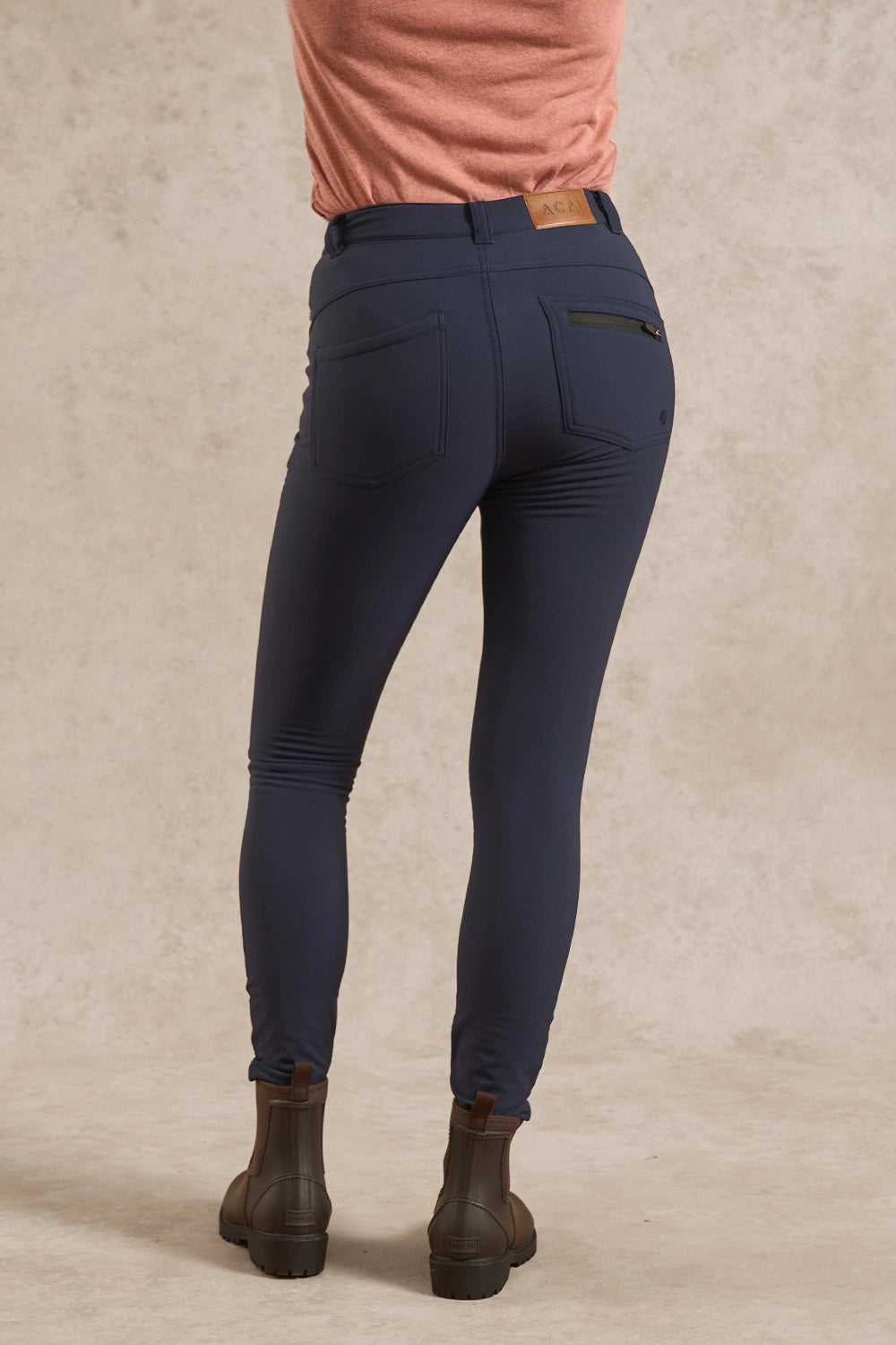 Rear view of a person wearing navy blue skinny equestrian riding pants with a zippered pocket on the back, paired with a peach top and brown leather ankle boots, against a textured beige studio backdrop.