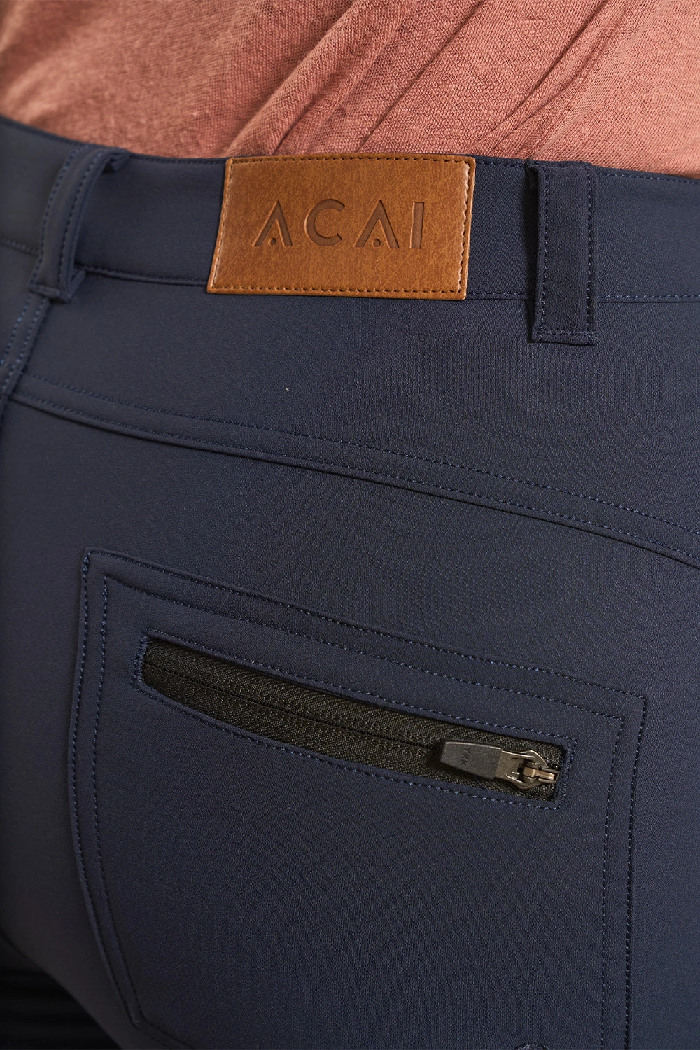 Close-up of navy blue pants with a brown leather patch at the waistband embossed with 'ACAI' and a zippered back pocket.