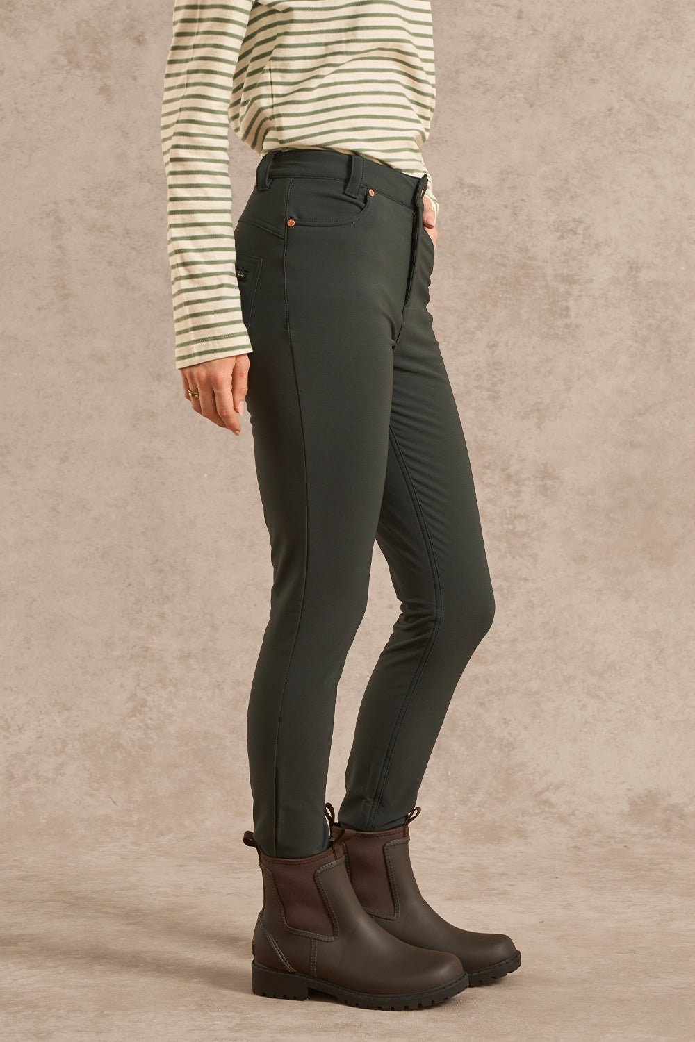 A person wearing slim-fit dark olive green riding breeches, a cream and green striped long-sleeve shirt, and dark brown Chelsea boots, standing against a textured beige studio backdrop.