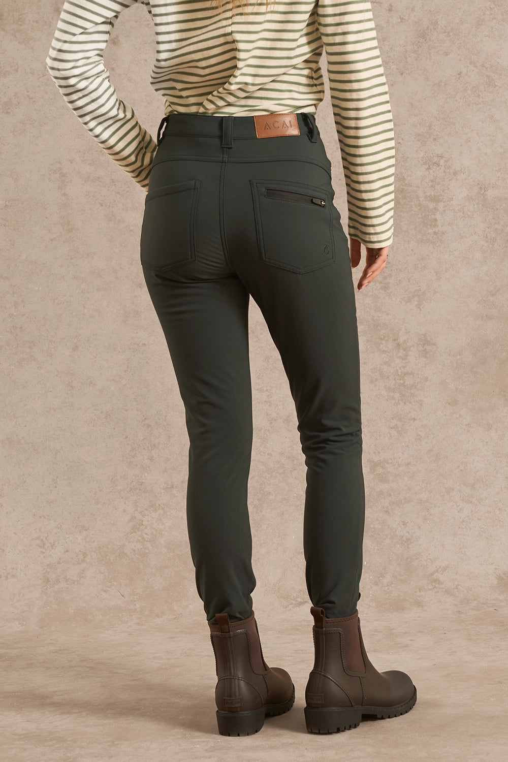 A person shown from the back wearing slim-fit dark olive green pants, a cream and brown striped long-sleeve top, and dark brown ankle boots, standing against a textured beige studio backdrop.