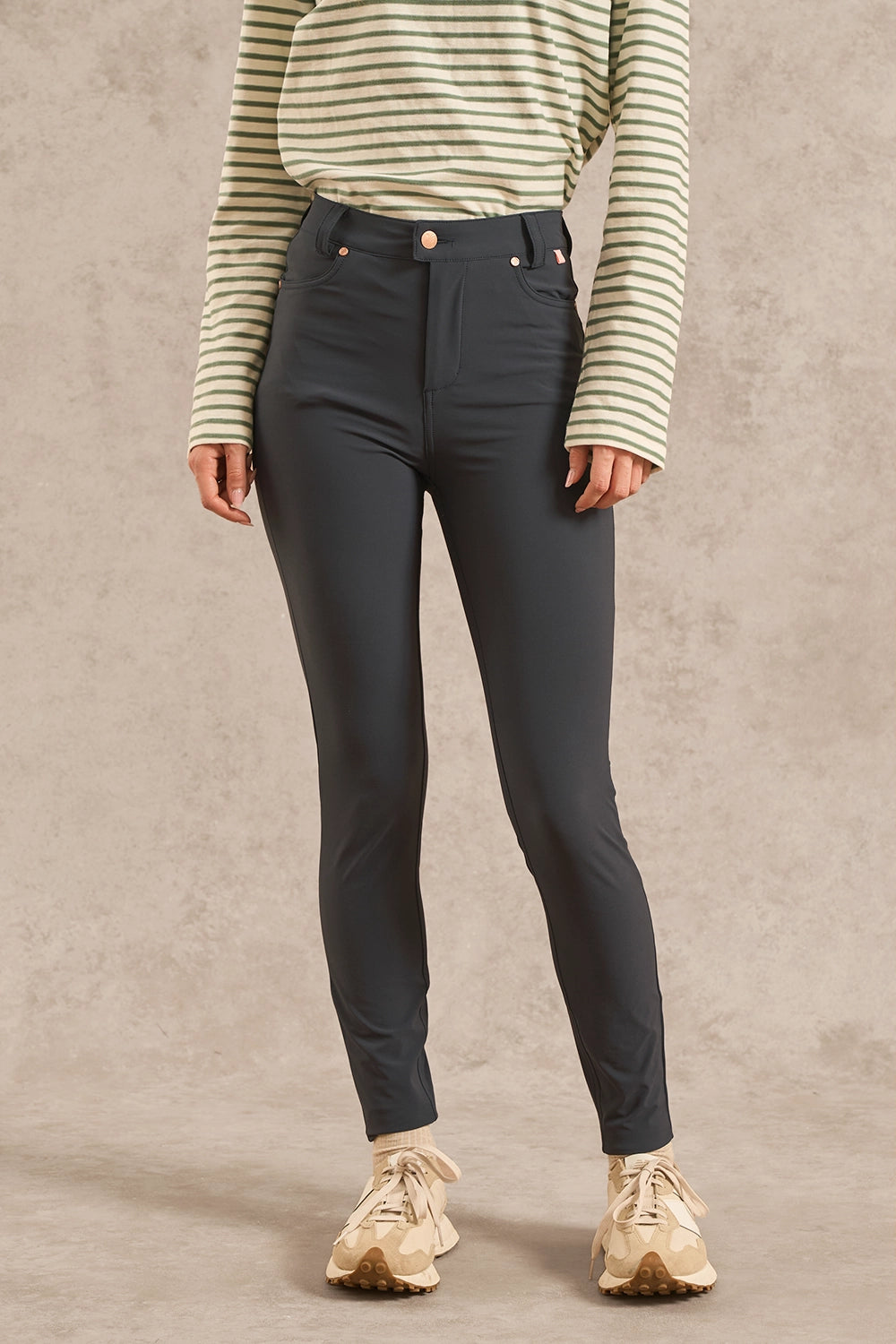 A model wearing dark charcoal gray slim-fit jeans, a green and cream striped long-sleeve shirt, and beige sneakers, standing against a textured beige studio backdrop.