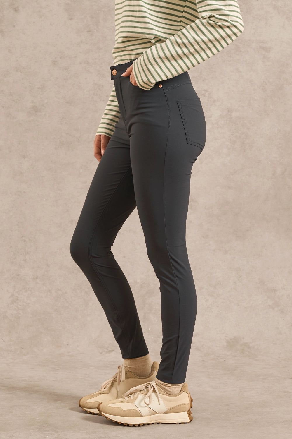 A person modeling slim-fit dark charcoal gray pants, paired with a green and white striped long-sleeve top and beige sneakers, standing against a textured beige studio backdrop.