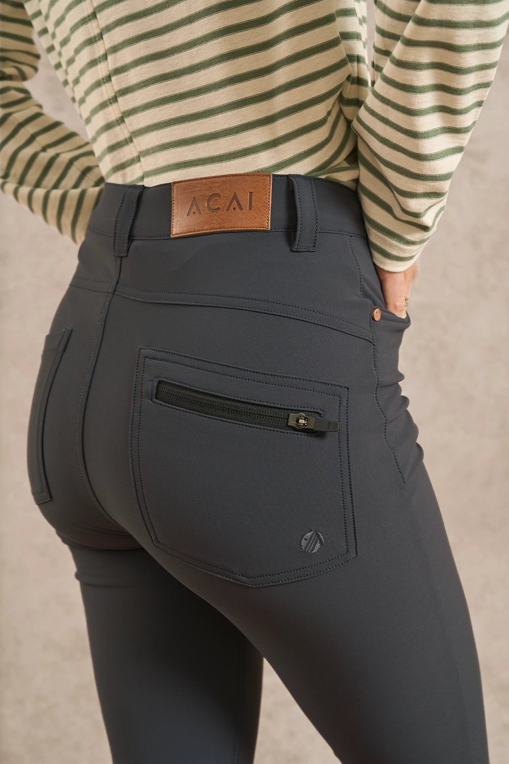 Close-up back view of a person wearing dark charcoal gray riding tights and a striped long-sleeve top. The tights have a zippered rear pocket and a small circular logo near the lower left leg.