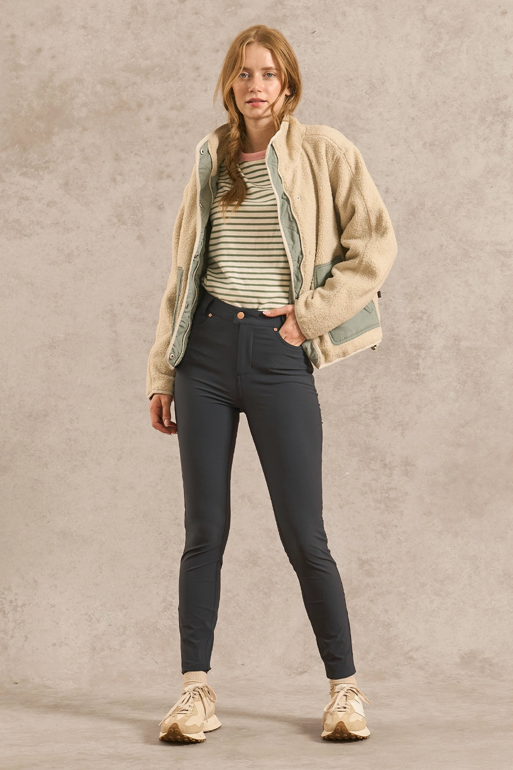 A woman stands against a textured beige studio backdrop wearing a cream sherpa jacket over a green and white striped shirt, dark gray skinny jeans, and beige sneakers.