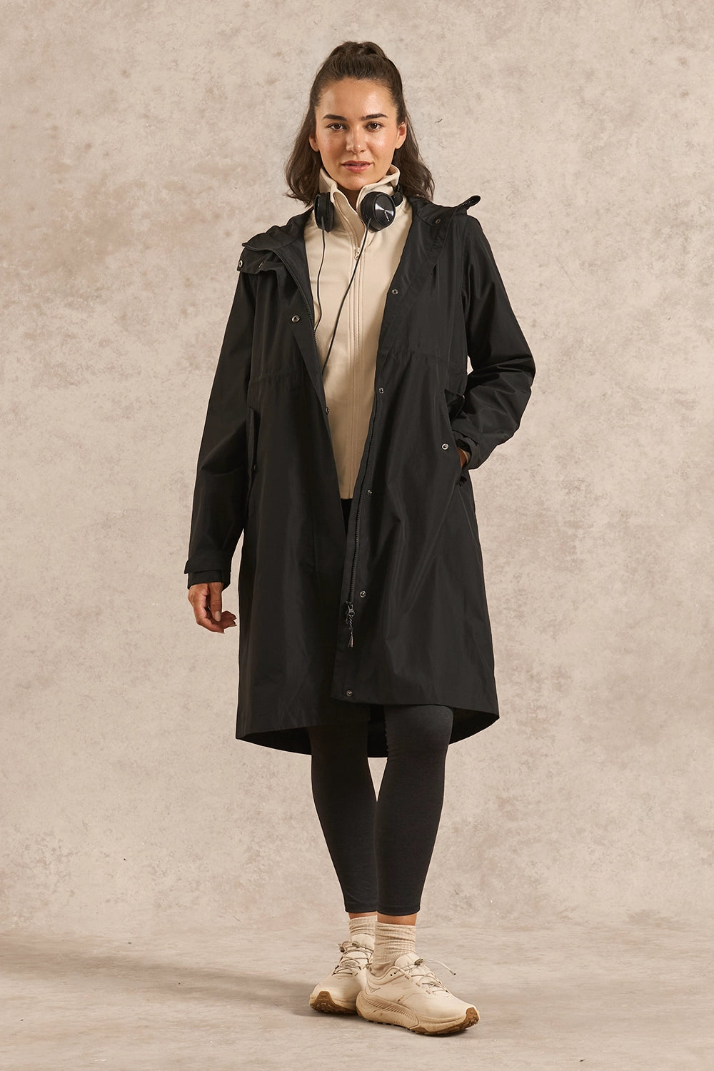 A woman modeling a long black hooded jacket over a cream top, black leggings, and beige sneakers, standing against a textured light beige studio backdrop.