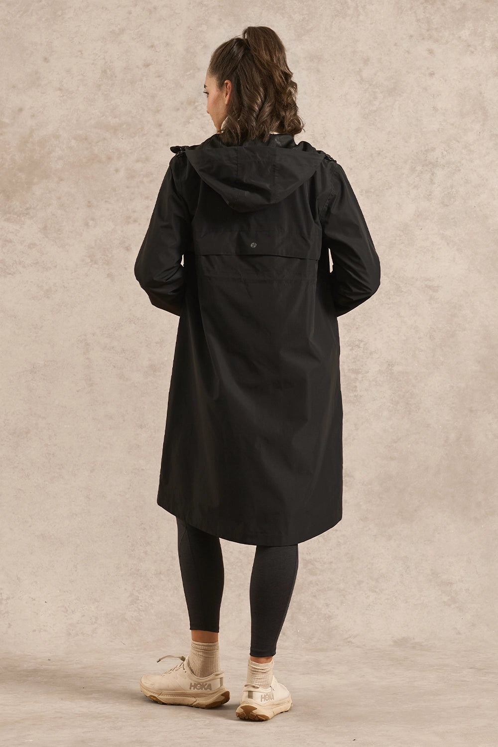 A woman viewed from behind wearing a black hooded coat over black leggings and cream sneakers, standing against a textured beige studio backdrop.