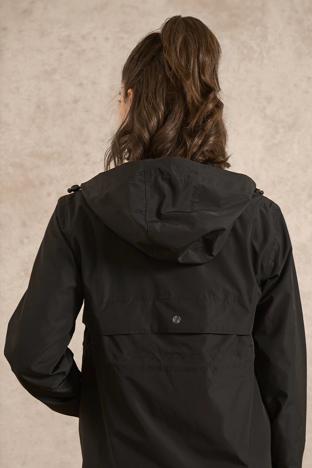 A woman viewed from behind wearing a black hooded jacket against a textured beige studio background.