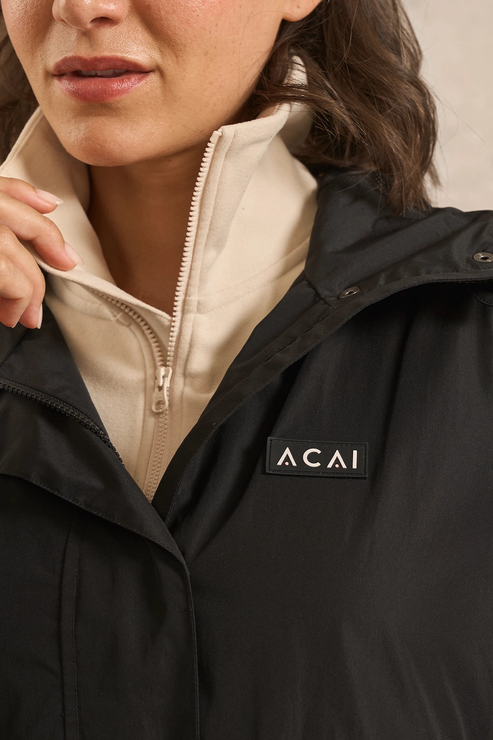 Close-up of a person wearing a black puffer jacket over a cream zip-up layer. The jacket has a small black rectangular patch on the chest with the brand name 'ACAI' in white.