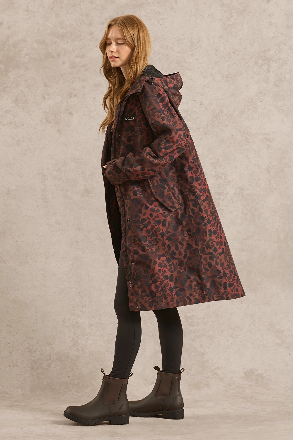 A woman wearing a long hooded coat with a dark red and black leopard print, black leggings, and dark brown Chelsea boots, standing against a textured beige studio backdrop.