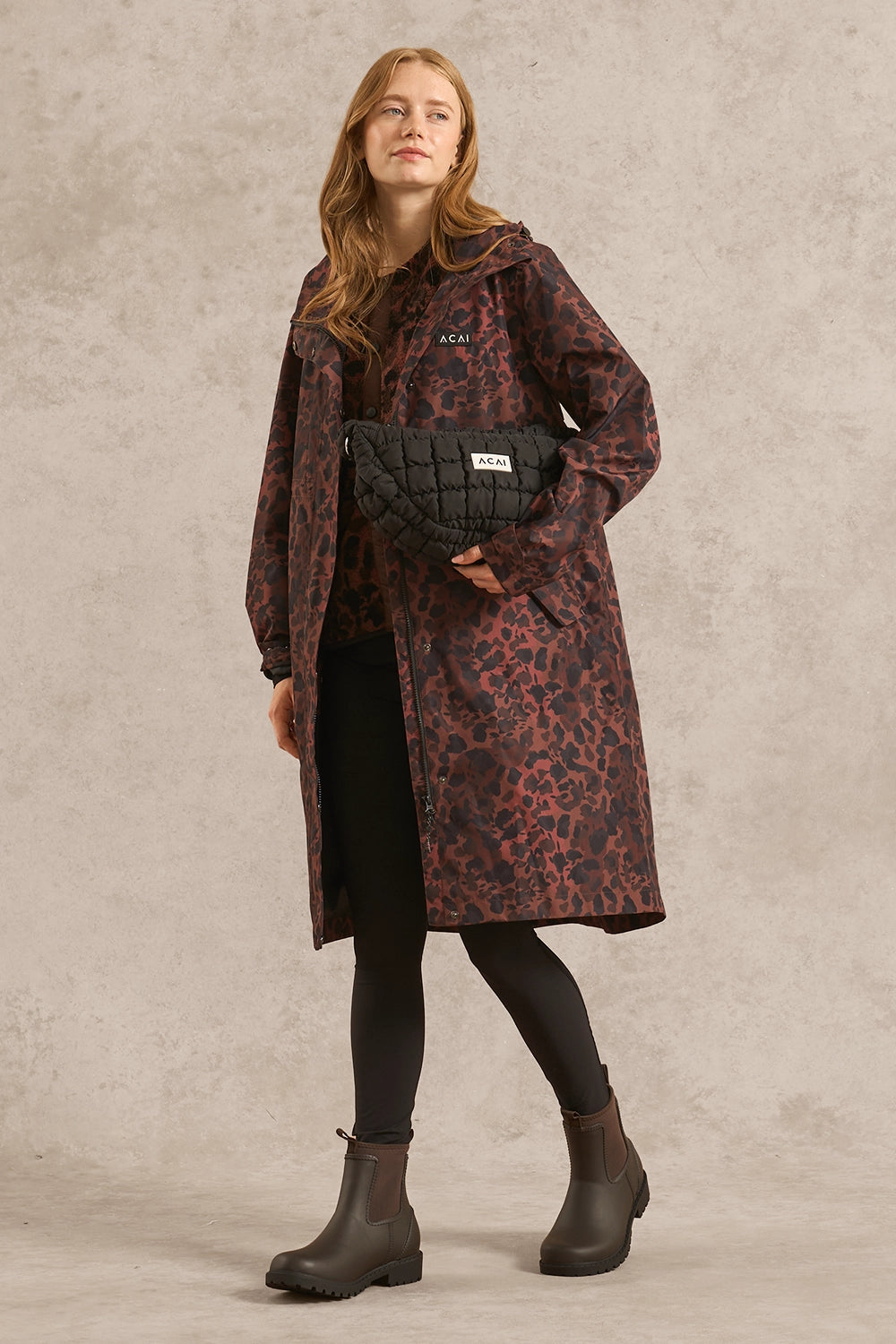 A woman models a long hooded coat with a dark red and black leopard print pattern, styled with a black quilted belt bag, black leggings, and dark brown rain boots against a textured gray studio backdrop.