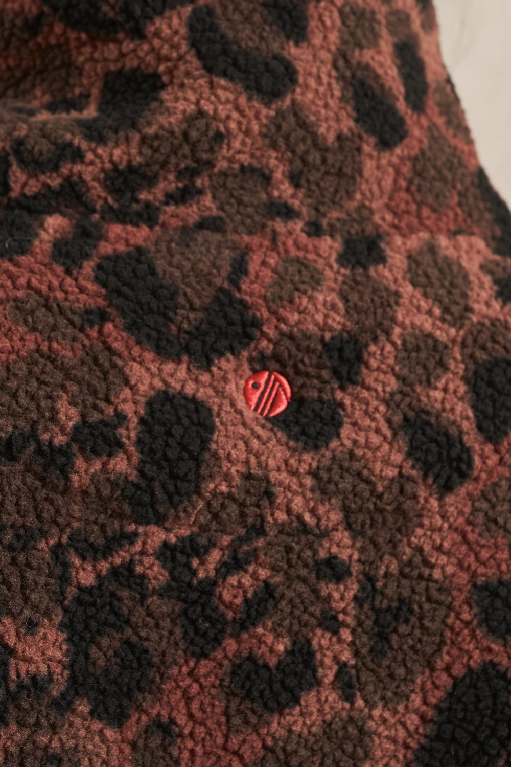 Close-up of a textured sherpa fabric with a leopard print pattern in dark brown and dusty rose, featuring a small red embroidered circular logo with white lettering.