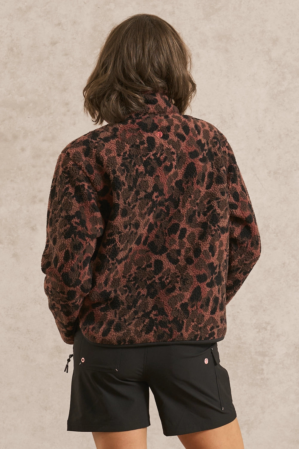 A model shown from the back wearing a brown and black leopard-print fleece jacket and black shorts, standing against a textured beige wall.