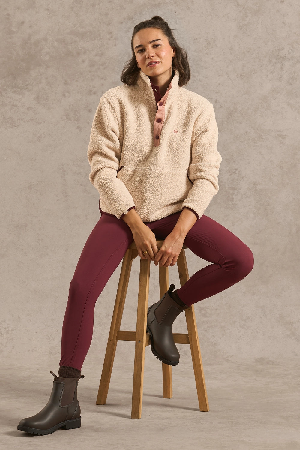 A woman sits on a wooden stool wearing a cream sherpa quarter-zip pullover, burgundy leggings, and dark gray Chelsea boots against a textured gray studio backdrop.