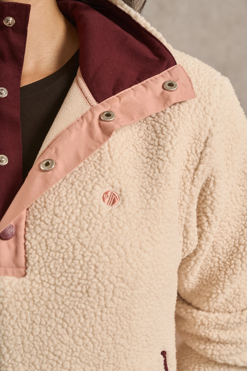 Close-up of a fleece jacket with a cream sherpa body, dusty pink snap placket, burgundy collar lining, and a small embroidered logo on the chest.