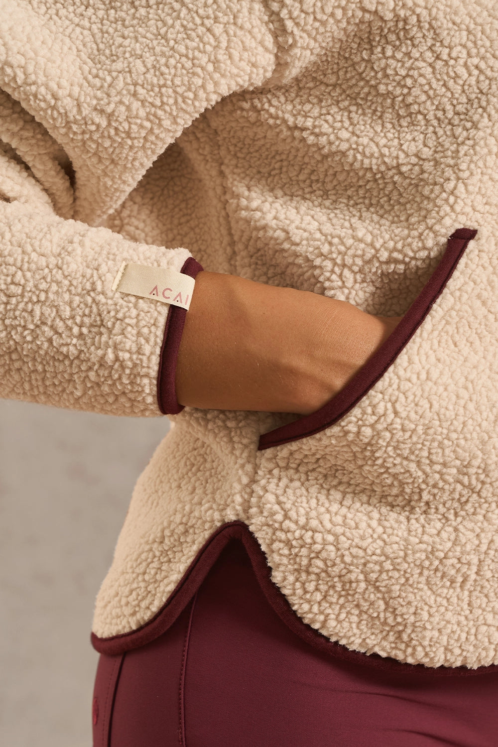 Close-up of a person wearing a cream sherpa fleece jacket with dark burgundy trim and a visible 'ACA' brand tag on the sleeve cuff, paired with burgundy pants.