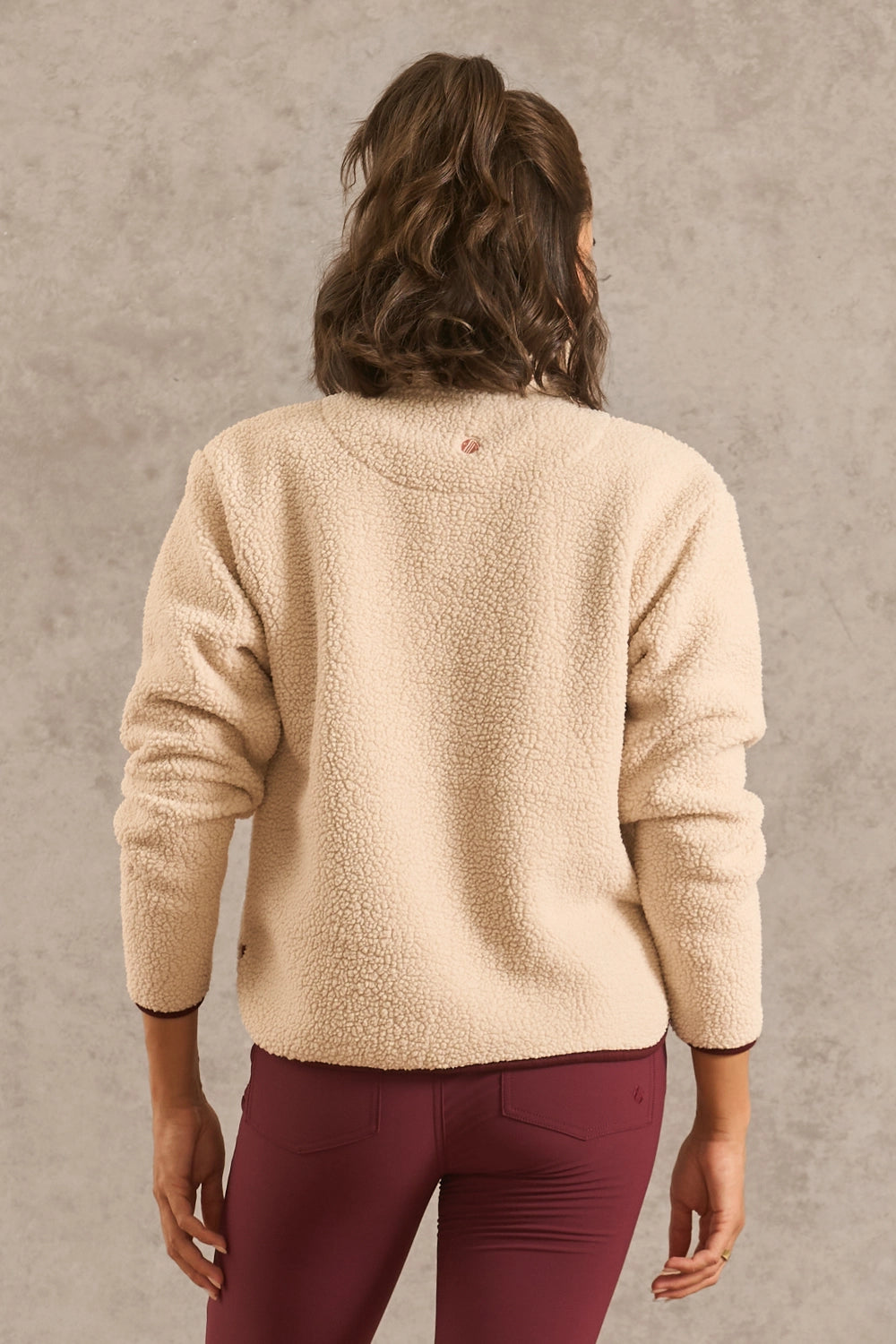 A woman viewed from behind wearing a cream sherpa fleece long-sleeve top and burgundy leggings, standing against a textured gray wall.
