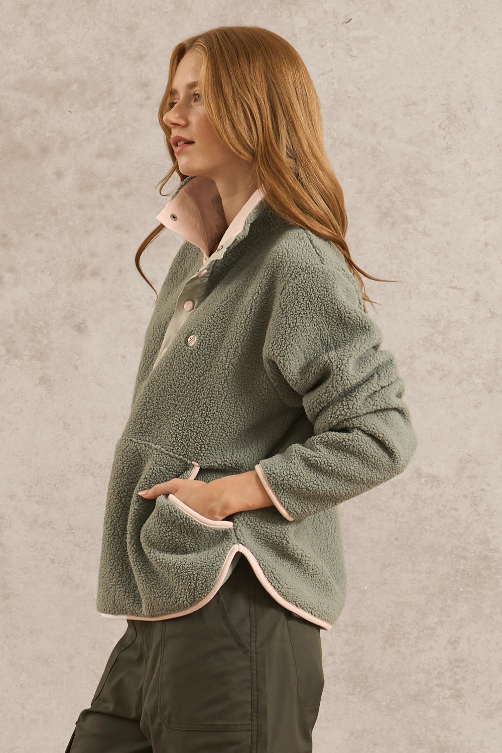 A woman wearing a sage green sherpa fleece button-up jacket with light pink lining, paired with dark olive pants, standing against a textured light gray wall.