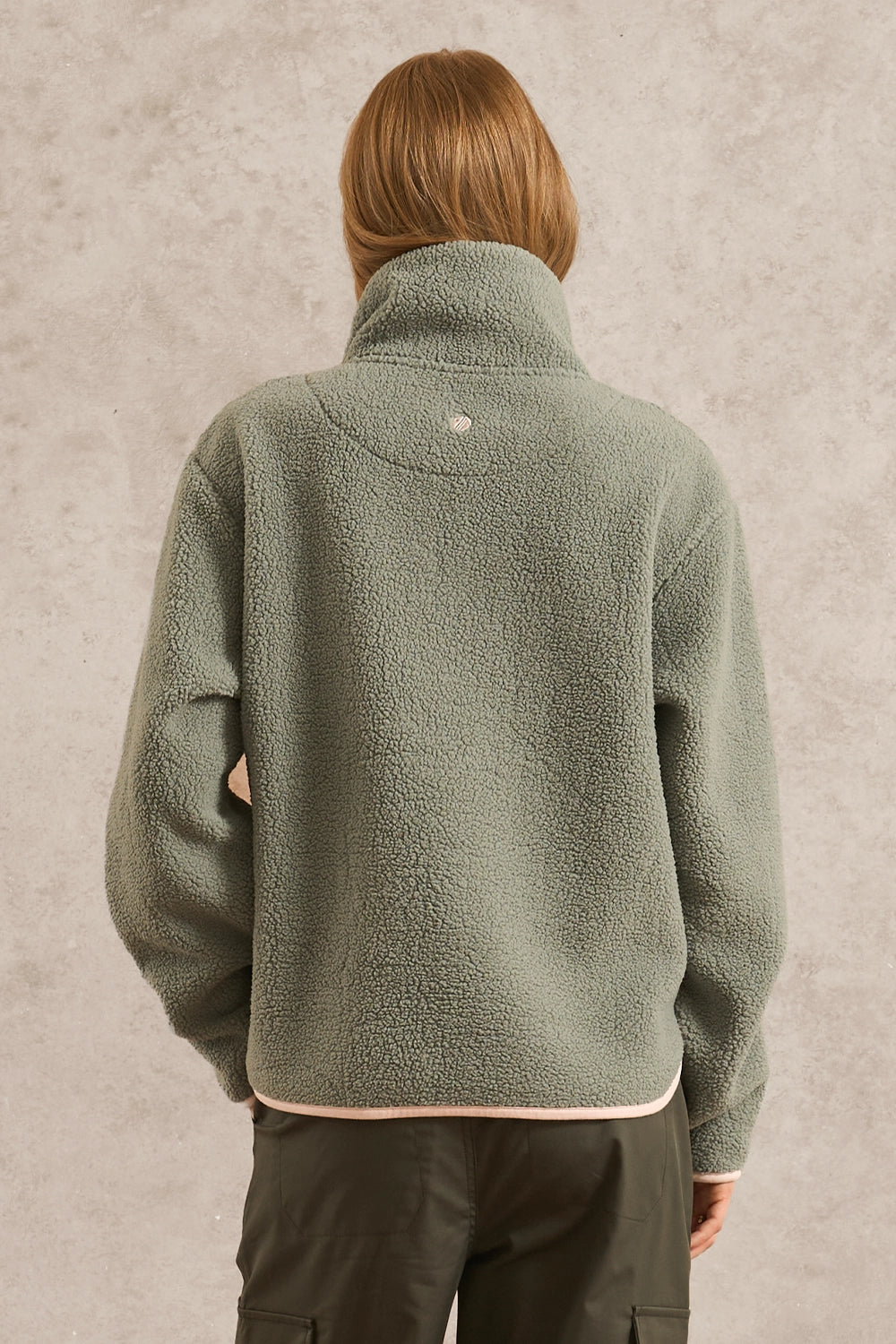 A person shown from the back wearing a sage green fleece pullover jacket with a high collar, paired with dark cargo pants, against a textured gray wall.