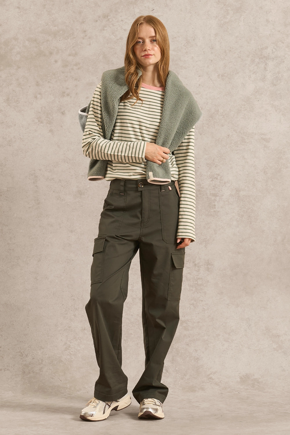 A woman stands against a textured light gray studio backdrop wearing a striped long-sleeve top, dark olive cargo pants, a sage green knit shawl draped over her shoulders, and white sneakers.