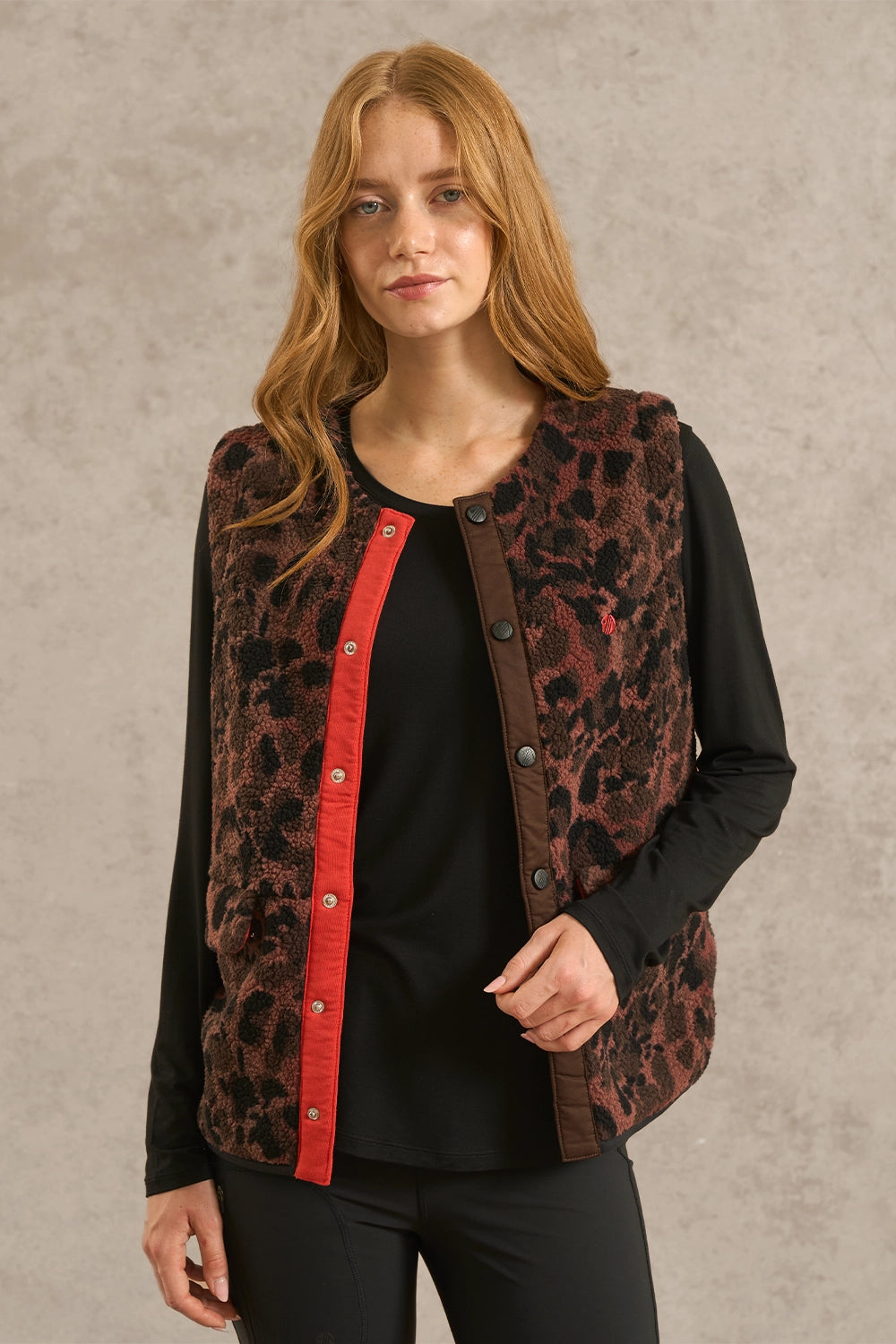 A woman modeling a sleeveless button-front vest with a brown and black leopard print pattern and a bright red trim down the front, worn over a black long-sleeve top against a textured gray studio background.
