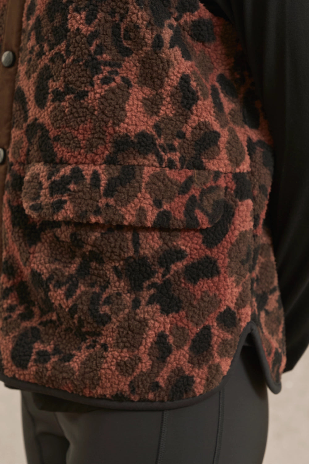 Close-up of a leopard-print fleece vest in rust red, brown, and black, worn over a black top and dark pants.