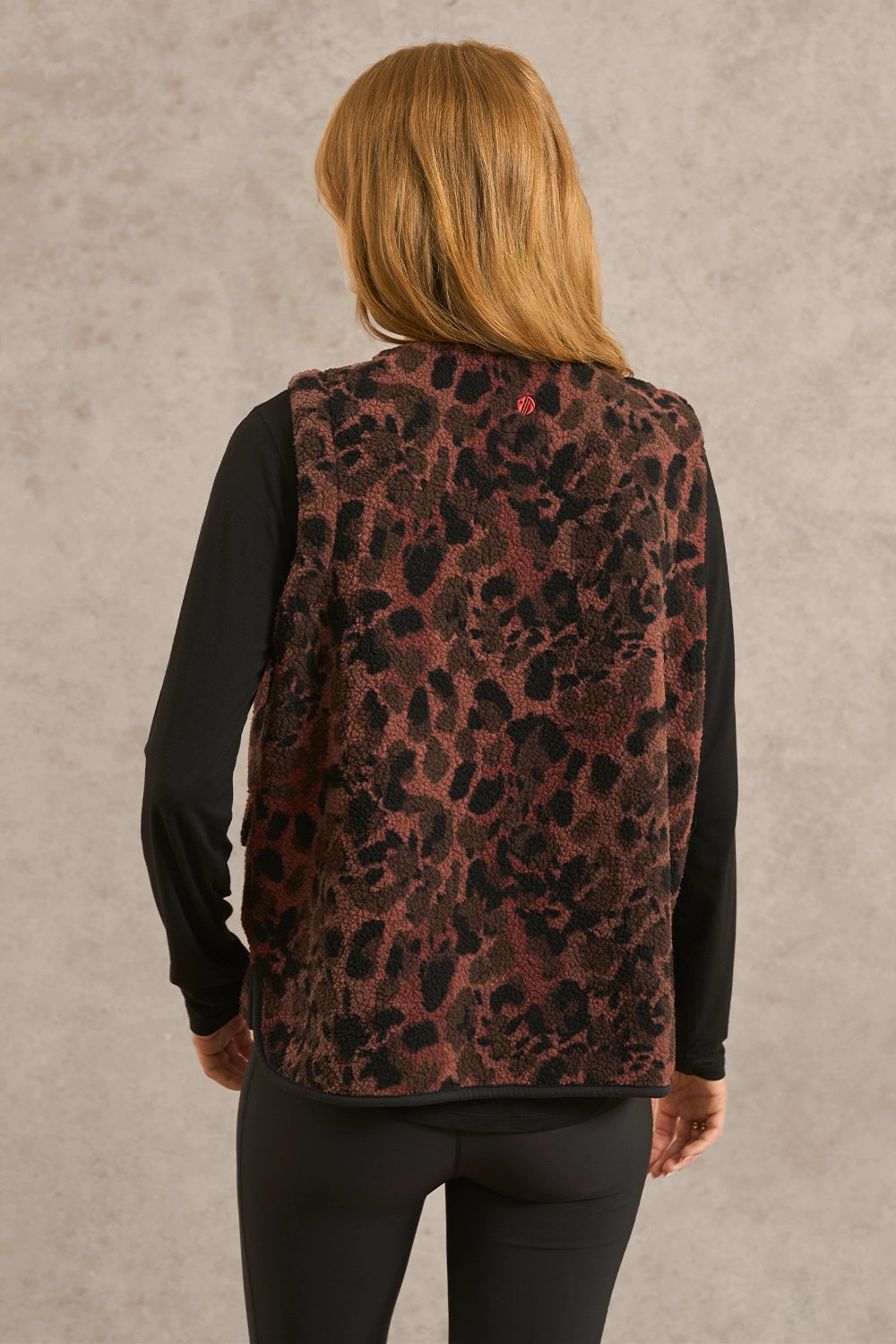 A woman viewed from behind wearing a sleeveless leopard-print vest over a black long-sleeve top and black leggings, standing against a textured light gray wall.