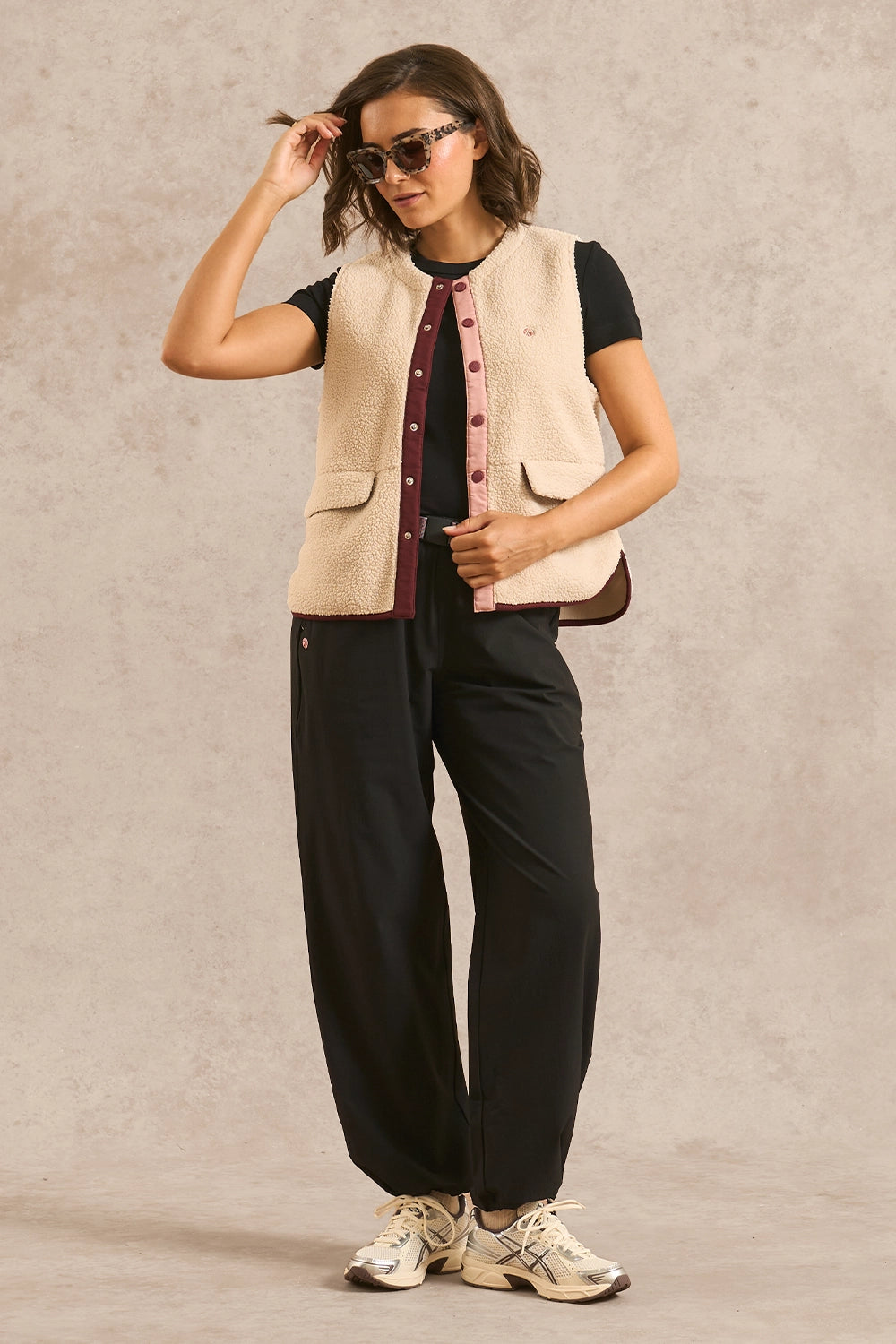 A woman models a cream-colored sherpa vest with pink buttons over a black t-shirt, black wide-leg trousers, and beige sneakers, standing against a textured beige studio backdrop.