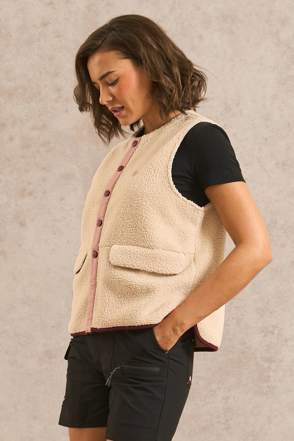 A woman wearing a cream sherpa fleece vest over a black t-shirt and black shorts, standing against a textured beige wall.