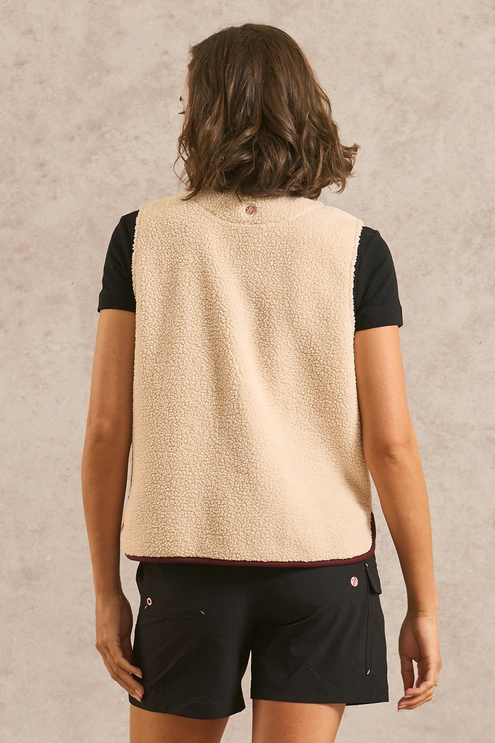 A woman shown from the back wearing a cream sherpa fleece vest over a black short-sleeve top and black shorts, standing against a textured beige wall.