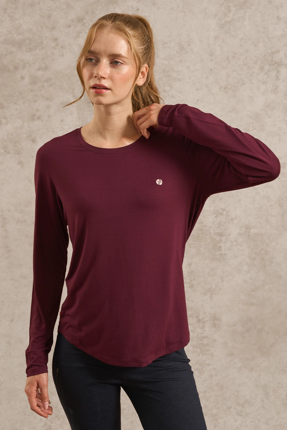 A woman wearing a long-sleeve crewneck top in deep burgundy with a small white circular logo on the left chest, paired with dark pants, set against a textured beige studio background.