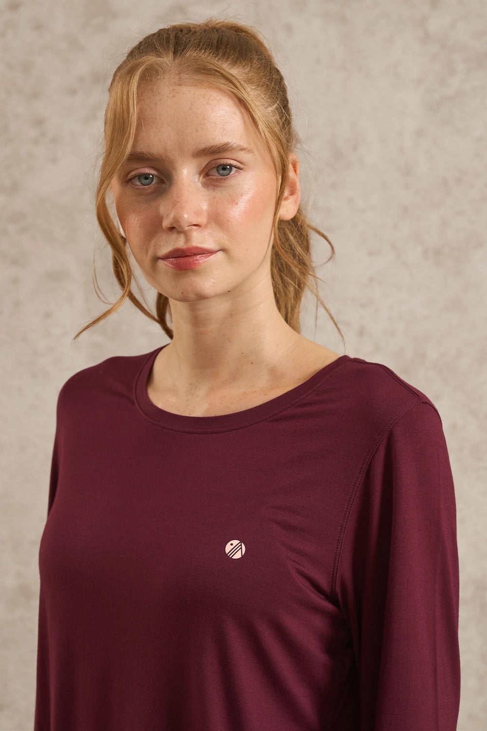 A woman with blonde hair tied back wears a long-sleeve crewneck top in a deep burgundy color, featuring a small circular metallic logo on the left chest. She is photographed against a textured, neutral beige background.