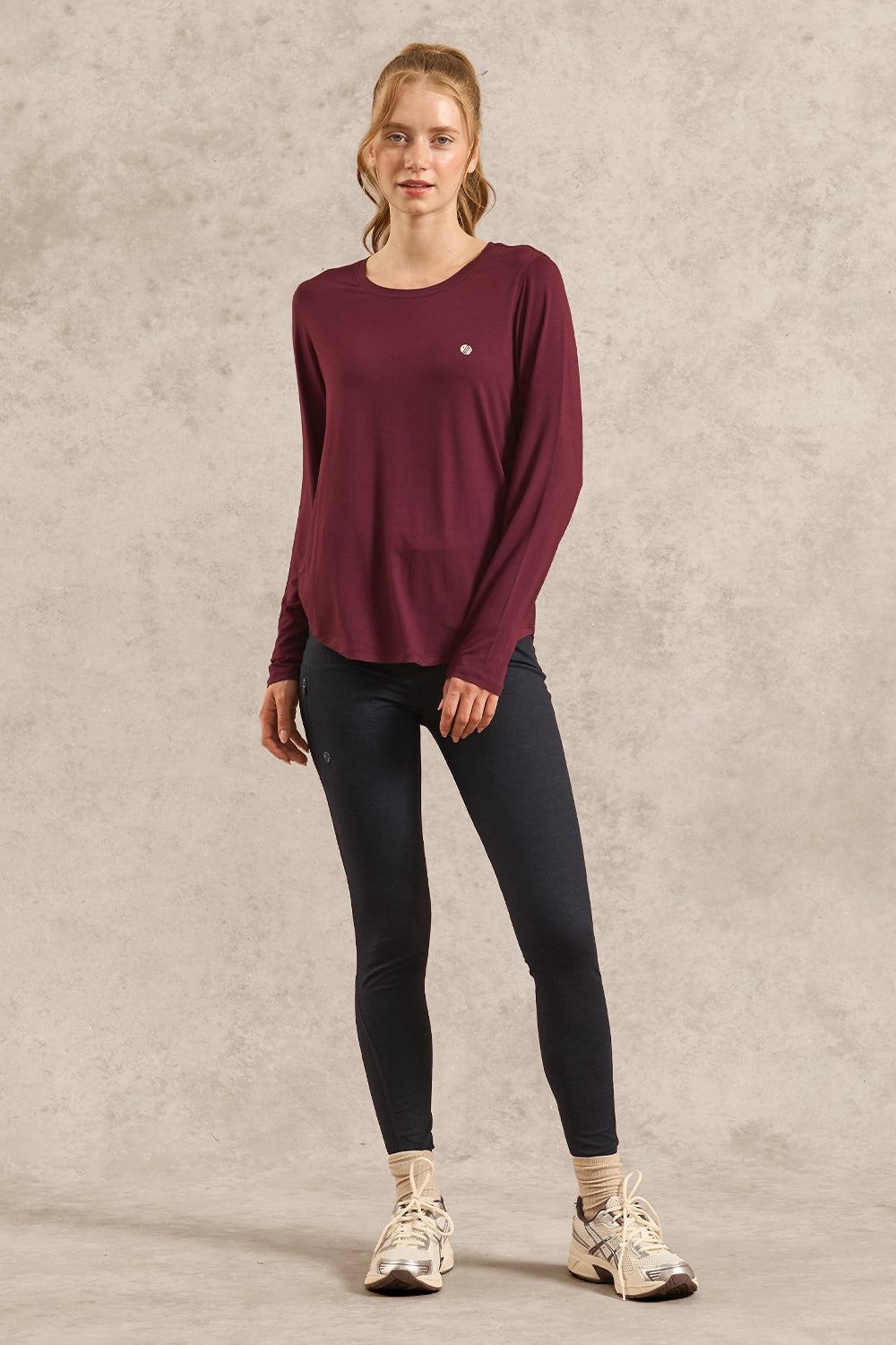 A woman modeling a long-sleeve crewneck top in burgundy, paired with dark navy leggings and white sneakers, standing against a textured beige studio backdrop.