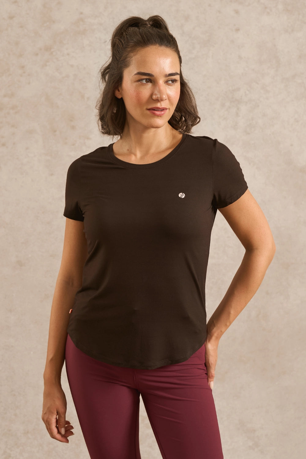 A woman modeling a dark brown short-sleeve athletic t-shirt with a small white logo on the left chest, paired with burgundy leggings, against a textured beige studio background.