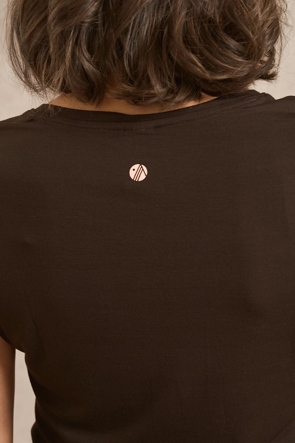 Close-up back view of a person wearing a dark brown short-sleeve t-shirt with a small circular metallic logo on the upper back.