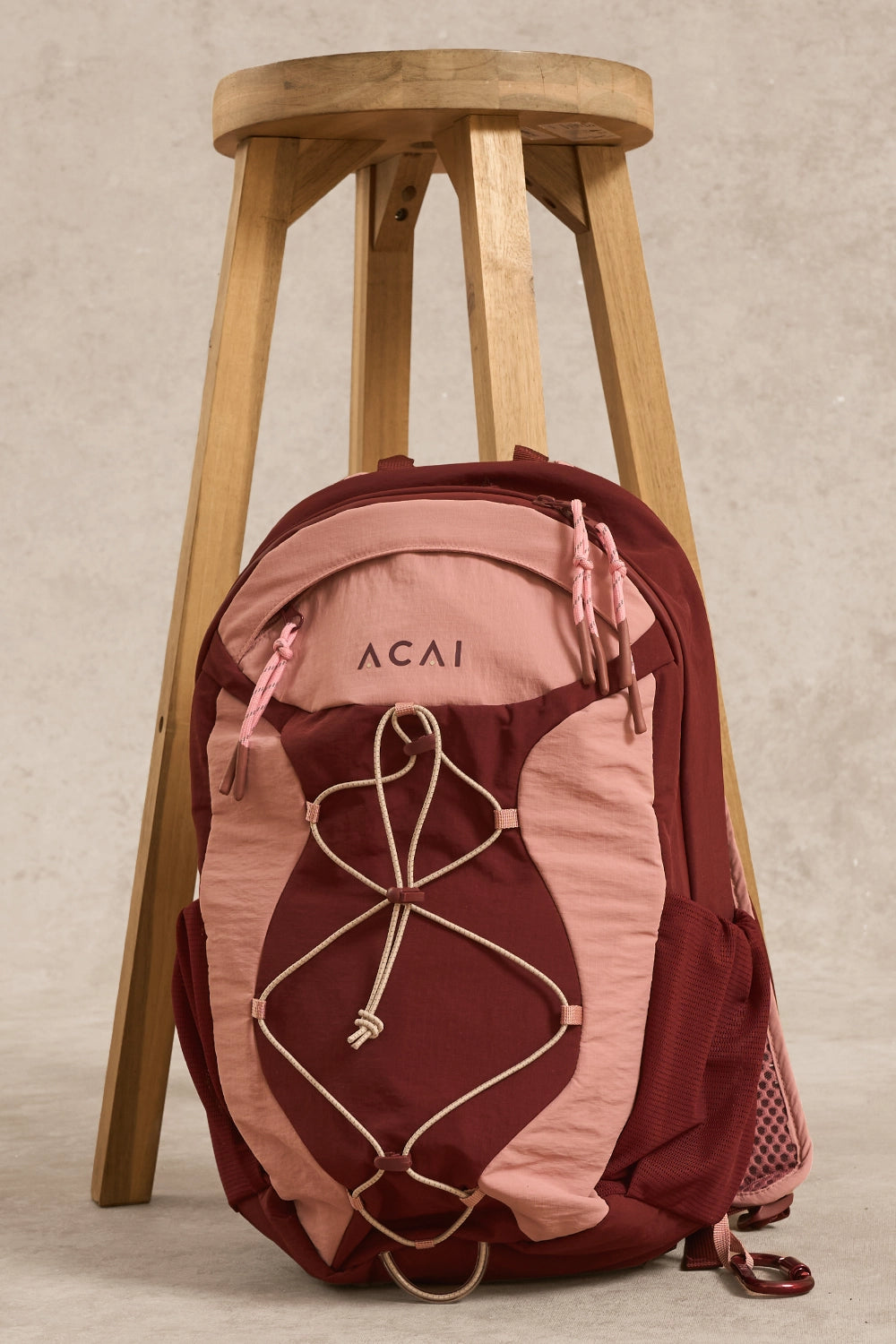 A pink and burgundy Acai backpack with beige webbing, leaning against a wooden stool on a textured beige background.