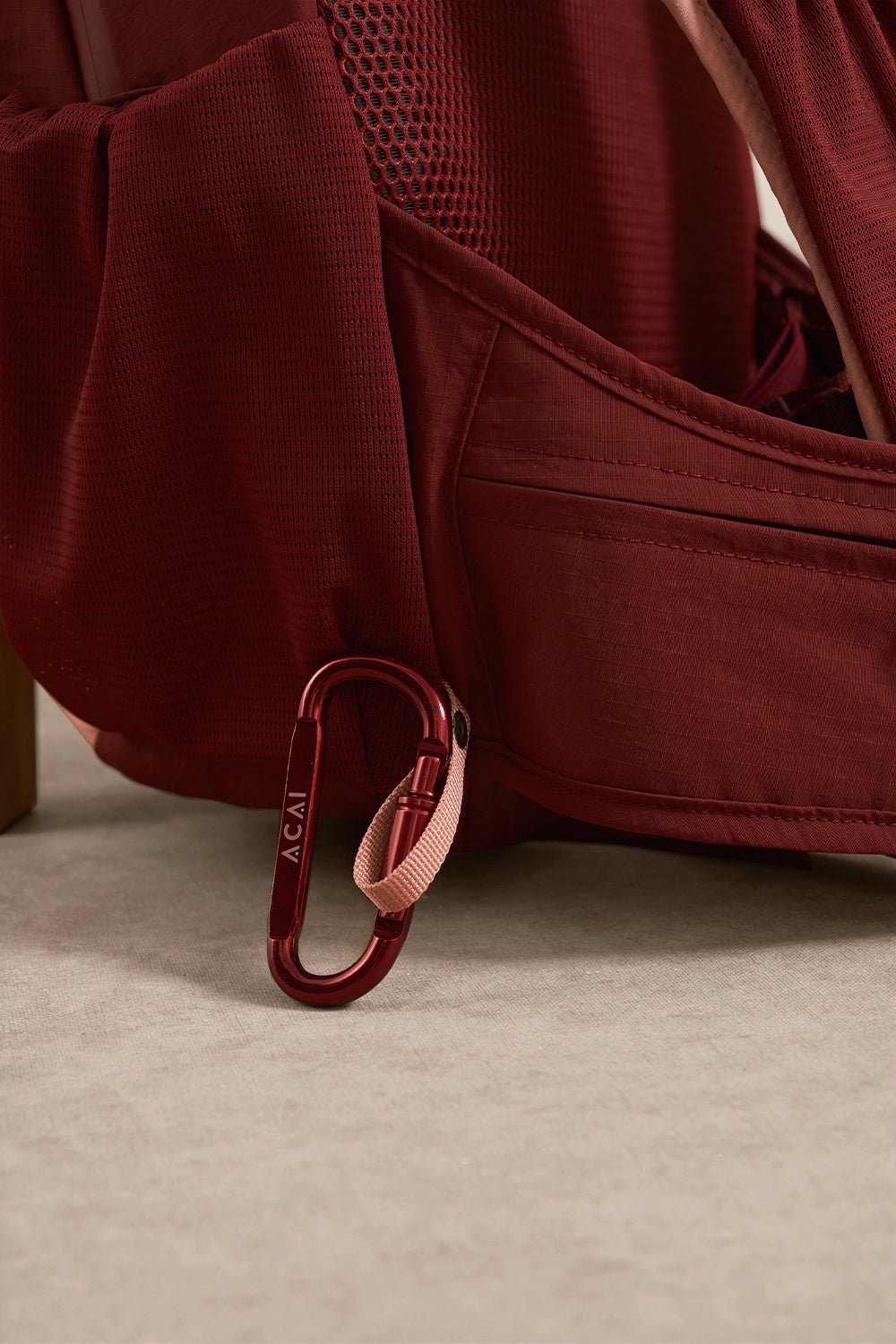 A close-up of a burgundy backpack with a matching carabiner and a light pink loop strap, resting on a light beige floor.