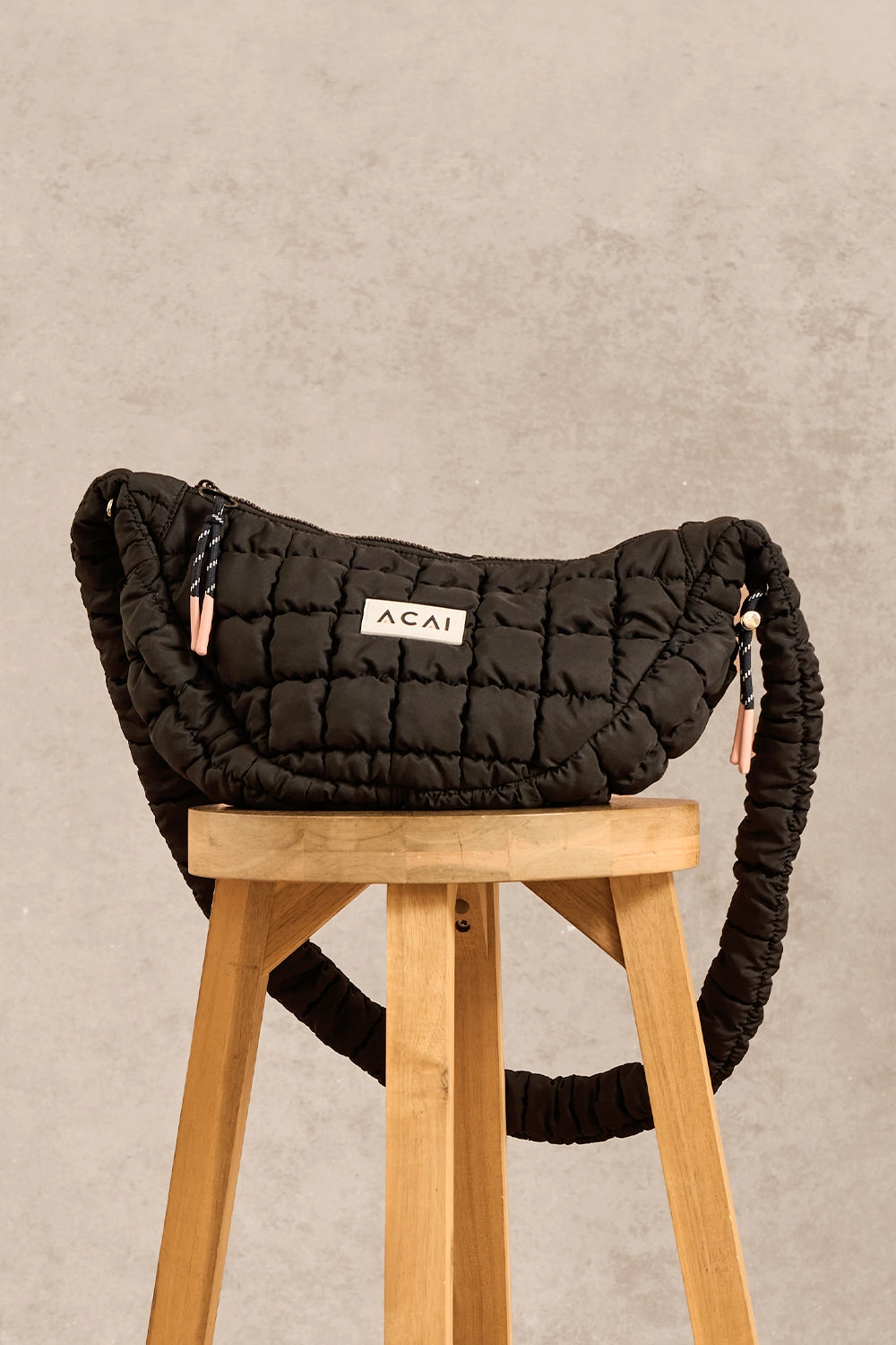 A black quilted puffer shoulder bag with a thick braided strap and a white 'ACAI' label, displayed on a wooden stool against a textured beige wall.