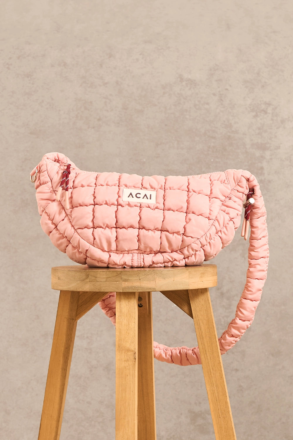 A pink quilted puffer shoulder bag with a white 'ACAI' label, displayed on a wooden stool against a textured beige wall.