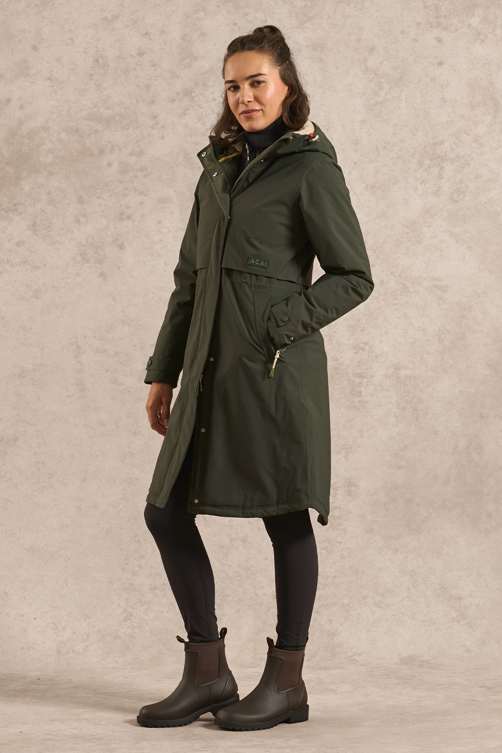 A woman modeling a mid-length olive green hooded parka coat with a waist belt, black tights, and dark brown ankle boots, standing against a textured light gray studio backdrop.