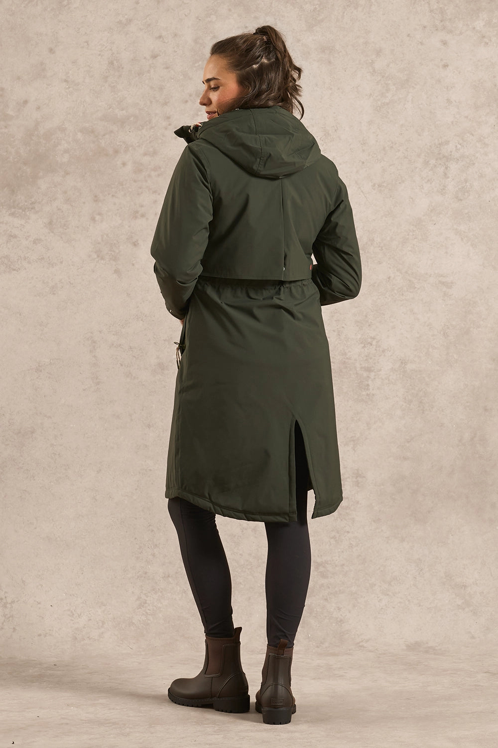 A woman wearing a dark olive green hooded parka coat, black leggings, and brown ankle boots, shown from the back against a textured beige studio backdrop.