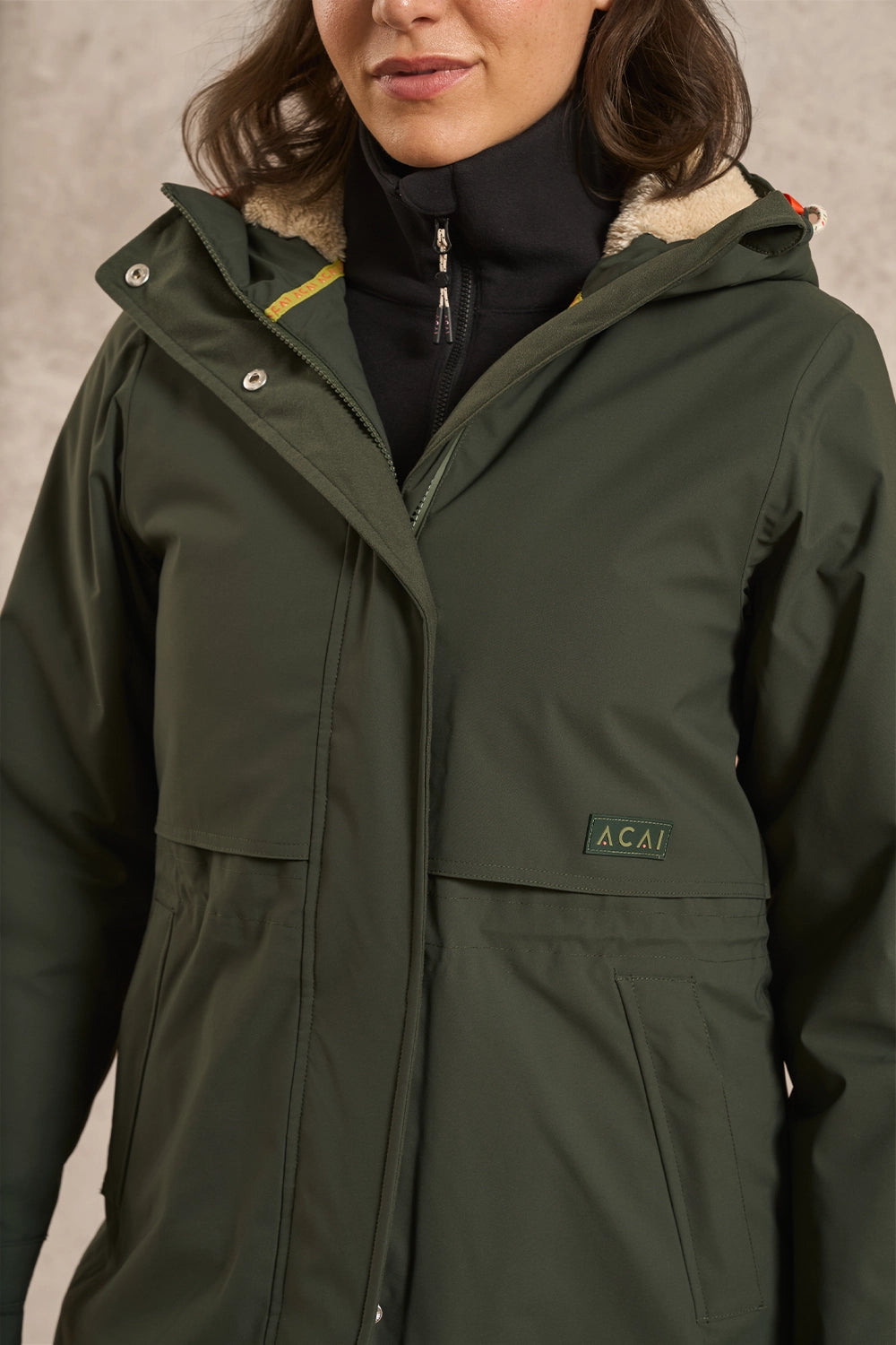A close-up of a woman wearing an olive green hooded jacket with a light beige sherpa lining and a small green Acai logo patch on the chest, layered over a black turtleneck.