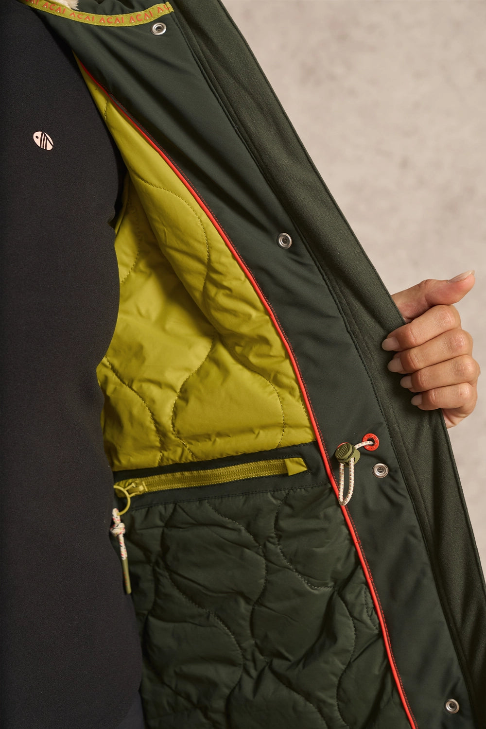 Close-up of a dark green quilted jacket with a bright yellow-green inner lining, red piping, and a hand holding the open flap.