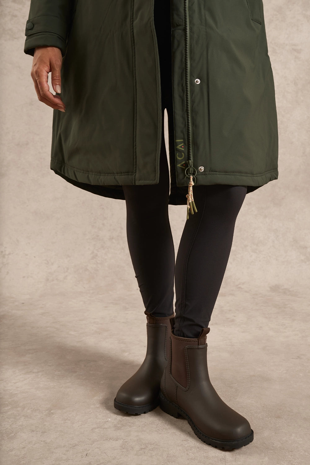 A person wearing an olive green mid-length parka coat over a black dress, paired with dark brown Chelsea-style boots, standing against a textured beige studio backdrop.