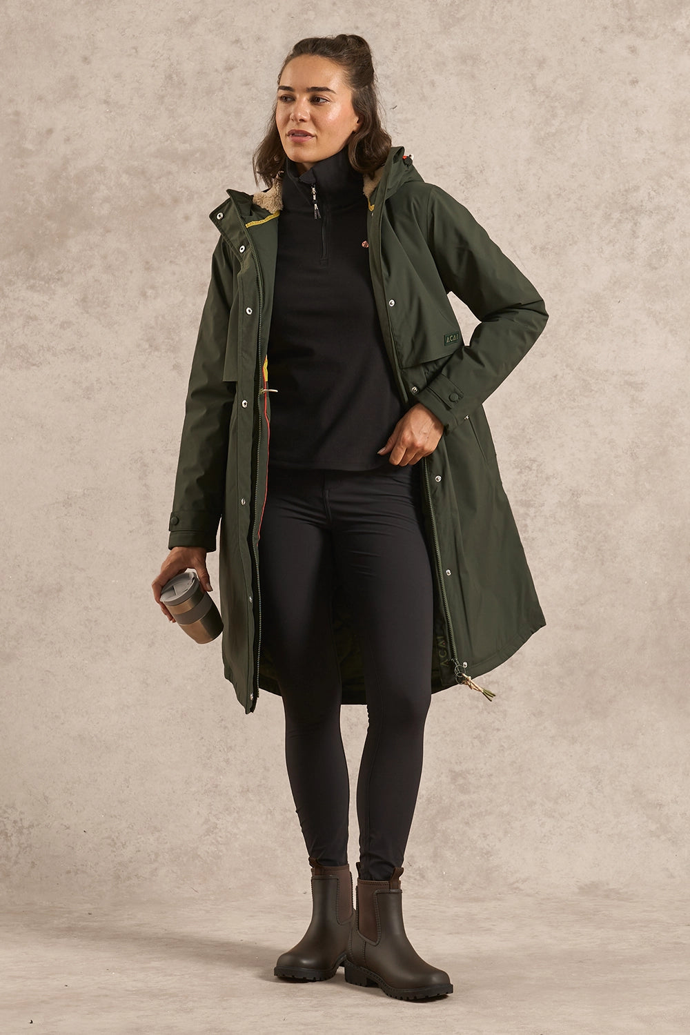 A woman models a mid-length olive green hooded raincoat over a black outfit, standing against a textured beige studio backdrop and holding a takeaway coffee cup.