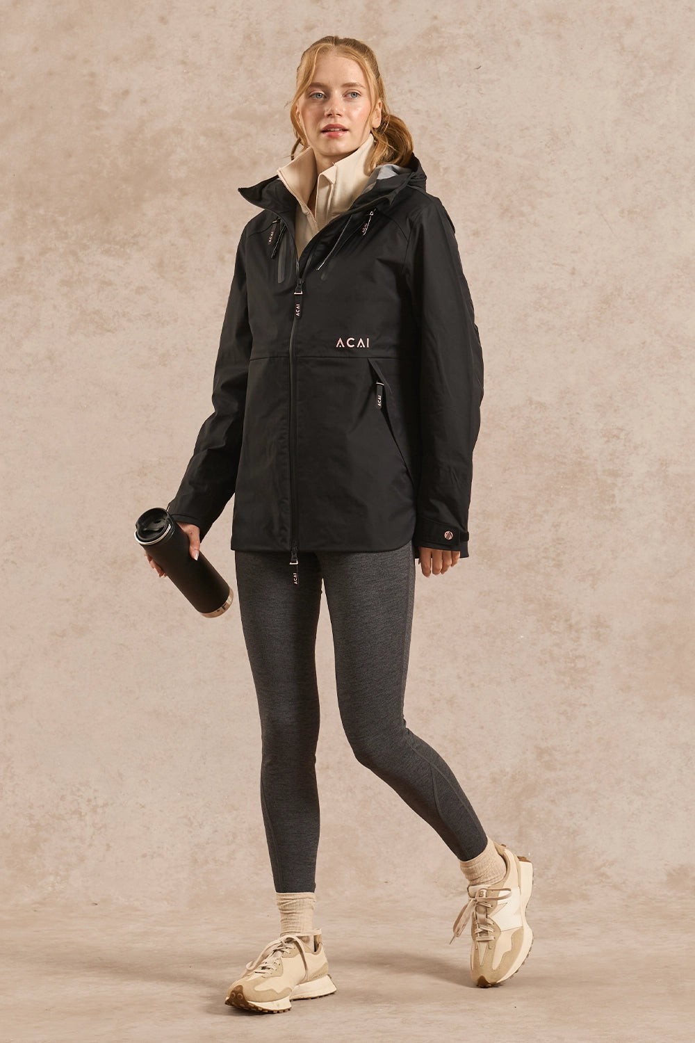A woman modeling a black hooded jacket with a beige collar, paired with dark gray leggings and beige sneakers, holding a black tumbler against a textured beige studio backdrop.