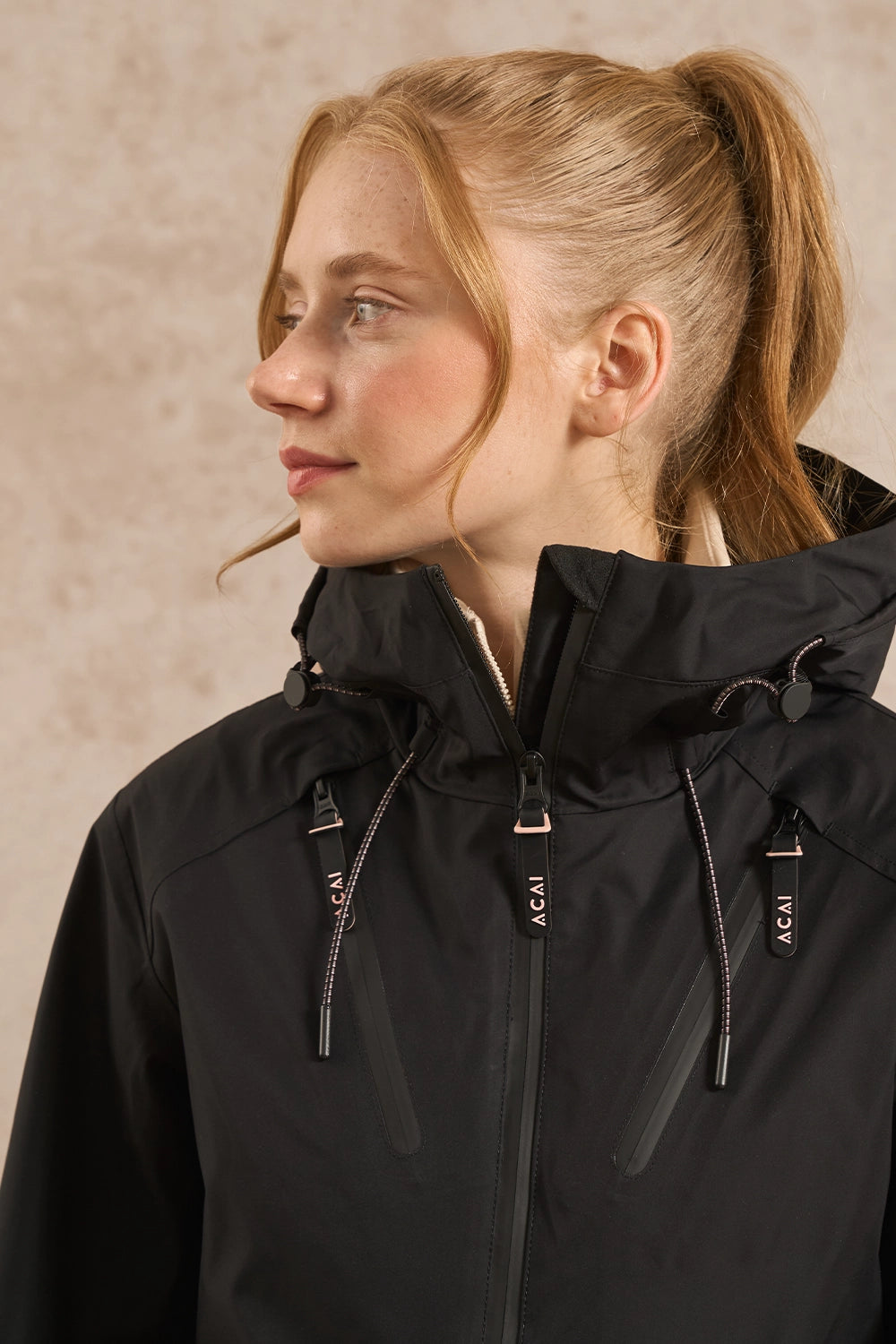 A woman with blonde hair in a ponytail wearing a black hooded jacket with silver zipper details and visible 'ACAI' branding, shown in a close-up profile view against a textured beige background.